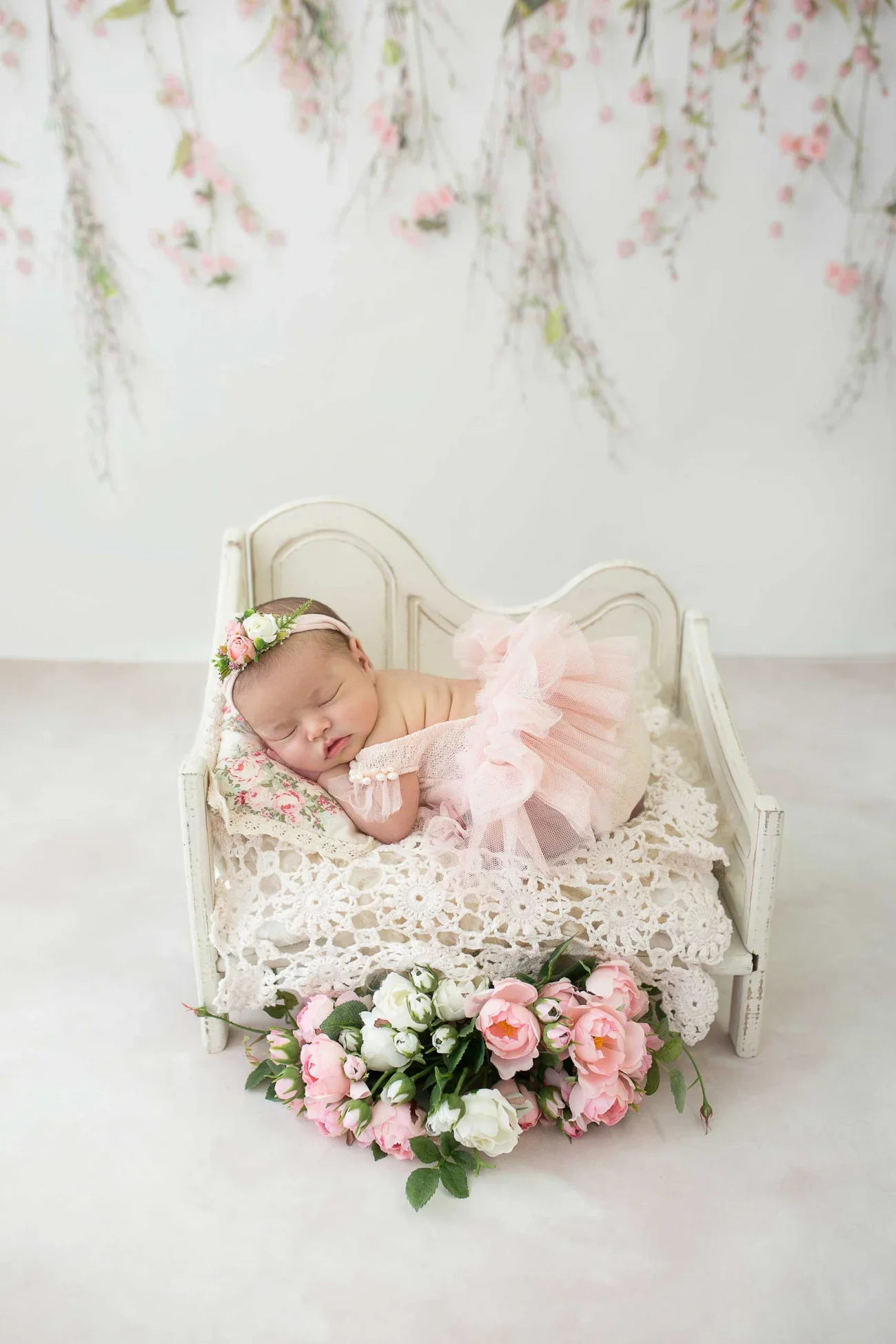 A sleeping baby girl lying on a small vintage white bed, dressed in a pink tutu and wearing a floral headband, surrounded by pink and white flowers, with pink flowers hanging on the wall in the background.
