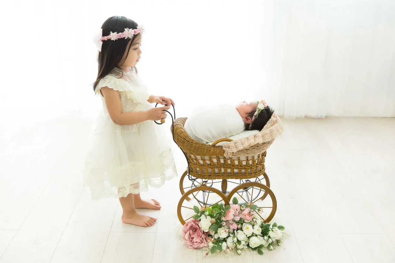 A young girl in a white lace dress and flower crown stares at a sleeping baby in a vintage wicker pram decorated with flowers in a bright, white room.