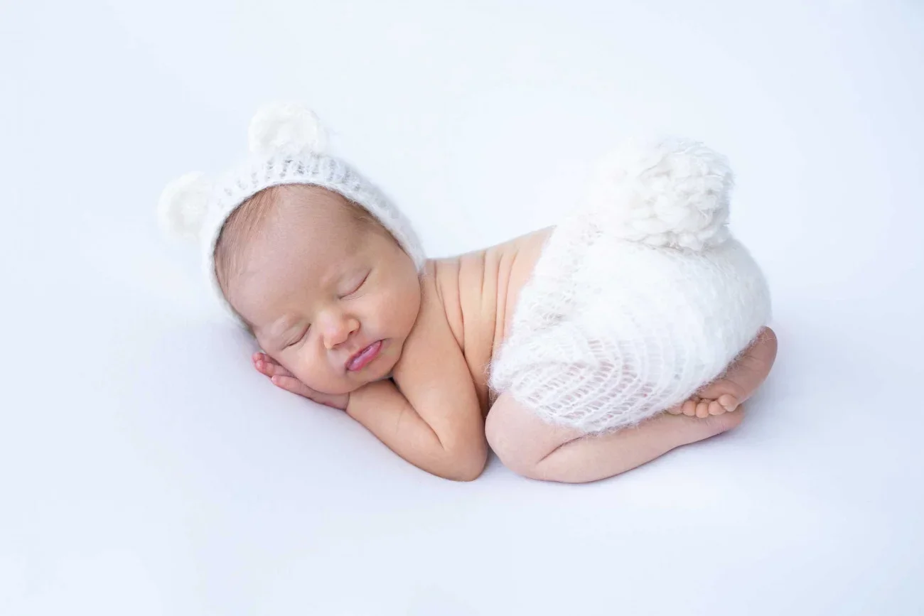 Adorable sleeping newborn baby wearing a white knit hat with pom-poms and a matching knit diaper cover, lying on a white surface.