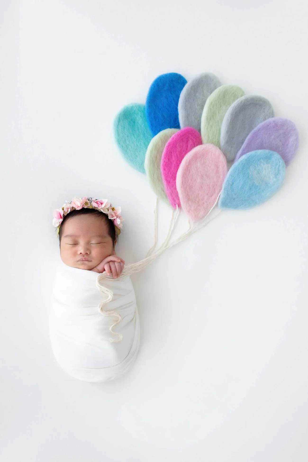 A sleeping baby wrapped in white cloth, with a flower headband, holding a string attached to colorful felt balloons resembling a cluster of balloons.