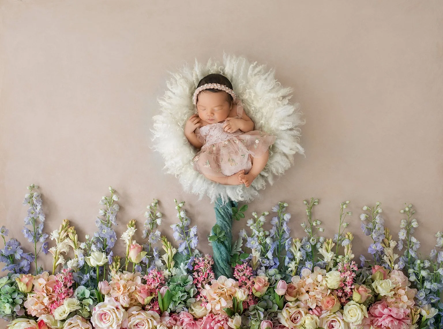 A newborn baby girl sleeping on a fluffy white cushion surrounded by an arrangement of pink, purple, and white flowers, with a floral headband and pink lace dress.