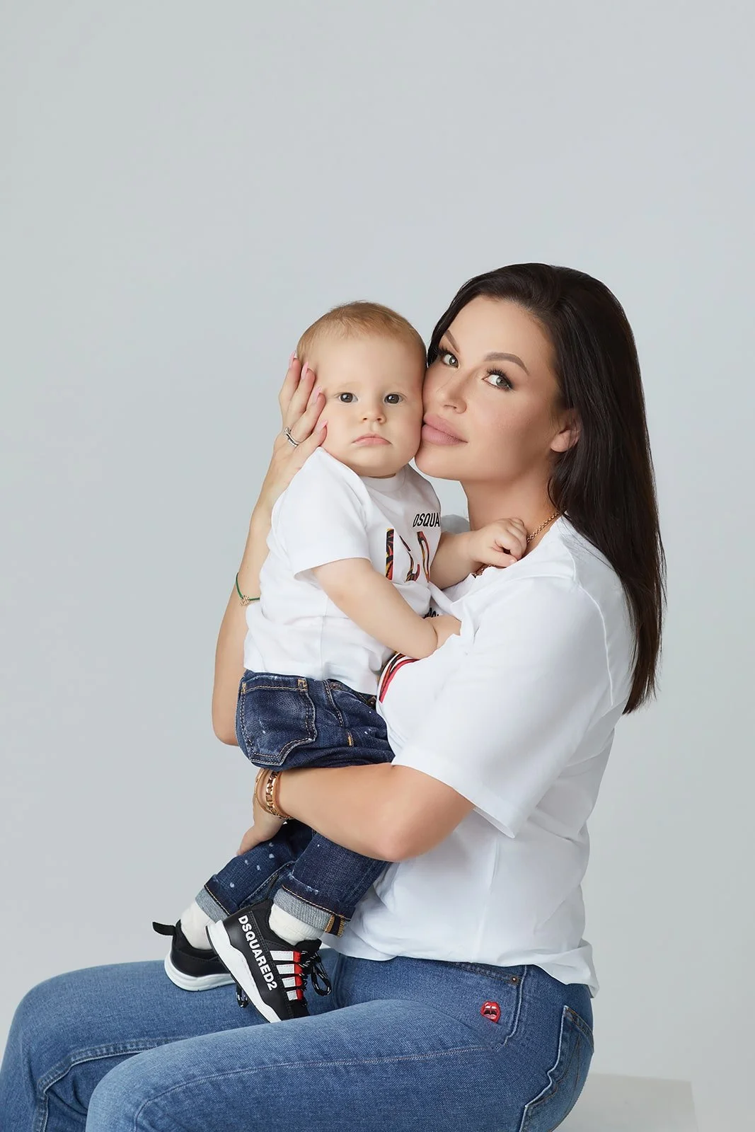 A woman with dark hair holding a young child with light hair, both wearing casual white shirts, against a plain light background.
