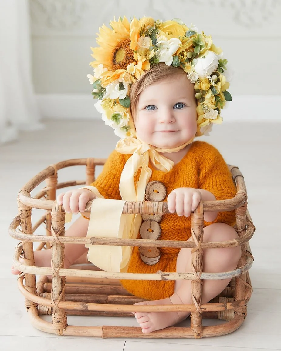 A baby girl wearing an orange knitted dress and a large sunflower and white flower floral crown, sitting inside a wicker basket, smiling at the camera.