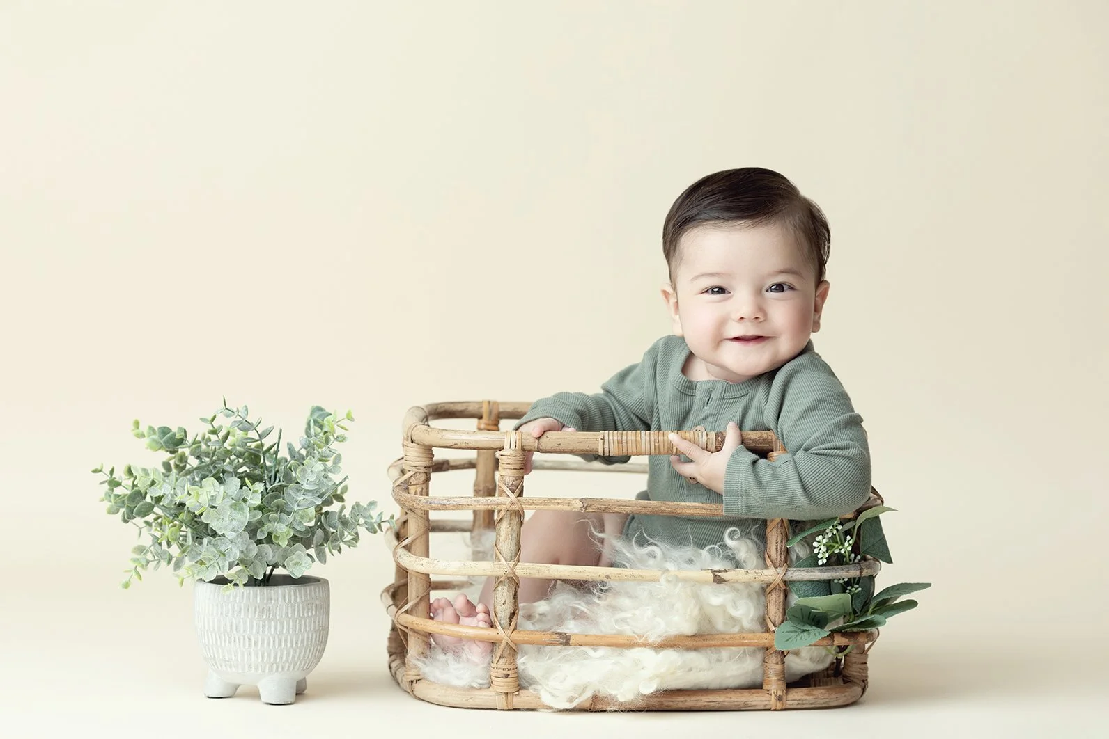 A smiling baby with dark hair, wearing a green shirt, sitting in a round woven basket with fluffy white fabric inside. Next to the basket is a white pot with green leafy plant. The background is plain and light-colored.
