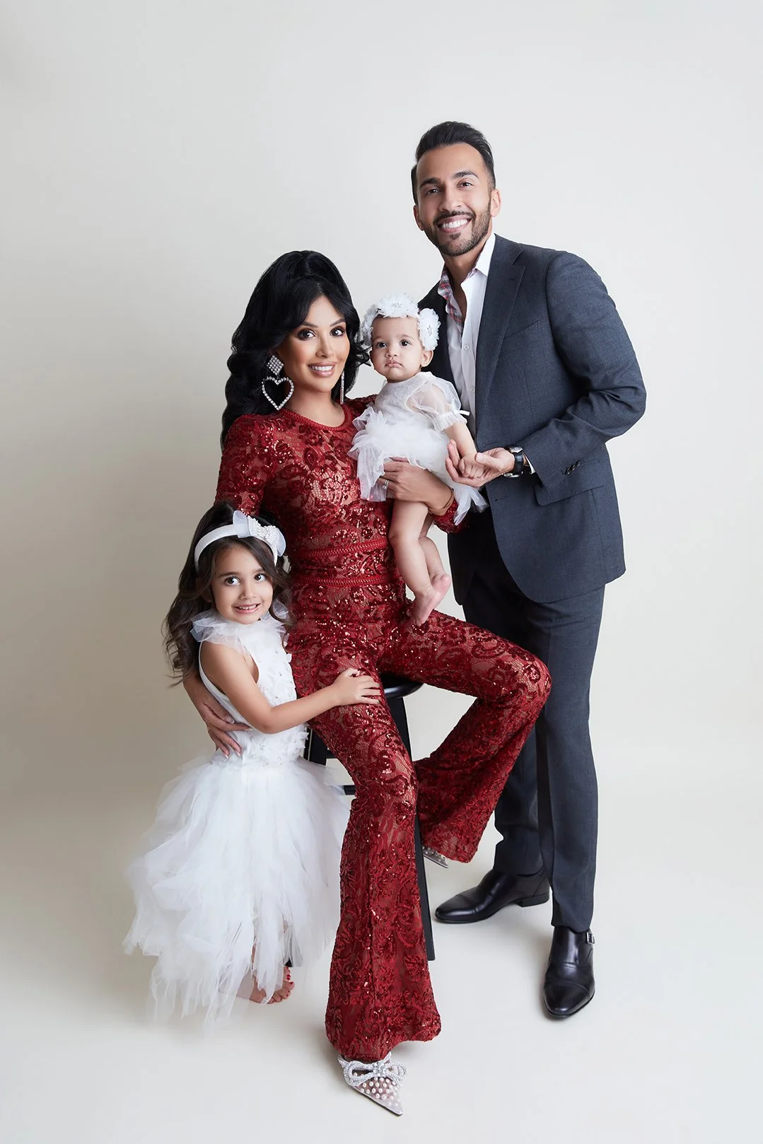 A family of four posing together, including a woman in a red lace jumpsuit, a man in a gray suit, and two young girls in white dresses, against a plain white background.