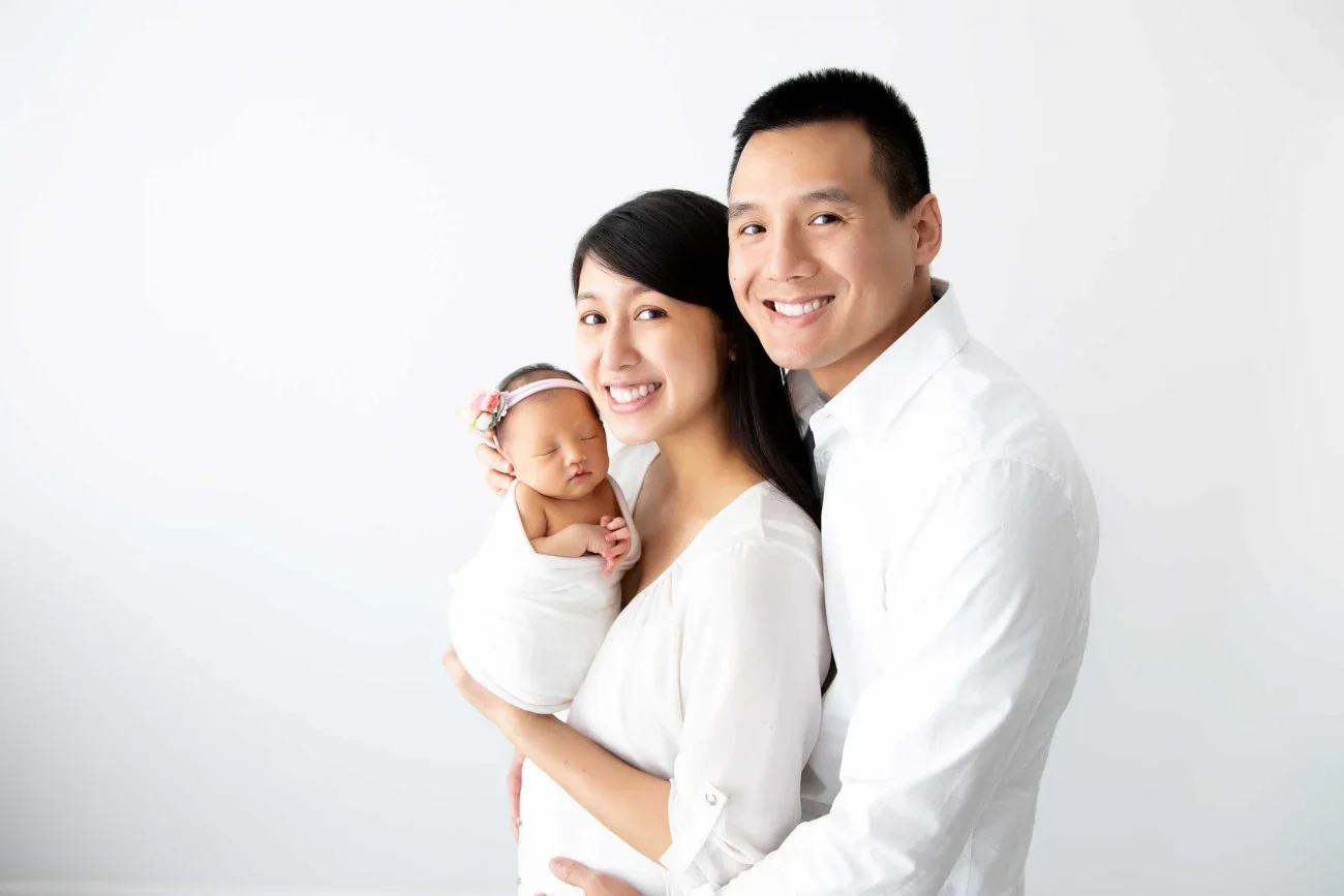 Family of three, Asian parents, holding their newborn daughter, smiling against a plain white background.