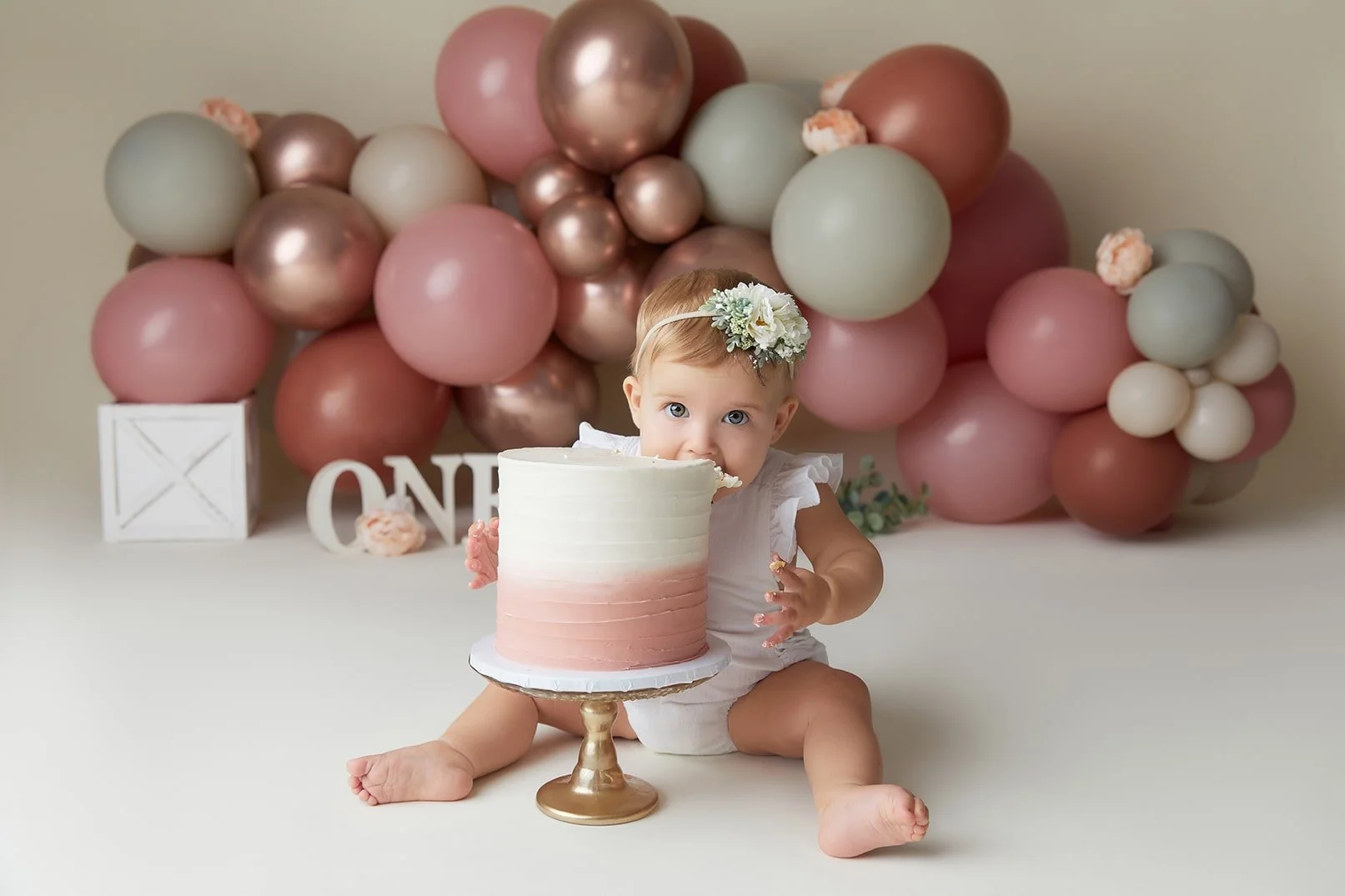 A young girl celebrating her first birthday, sitting on the floor with a partially eaten cake in front of her, with a backdrop of pink, rose gold, white, and muted green balloons. She is wearing a floral headband and a white outfit.