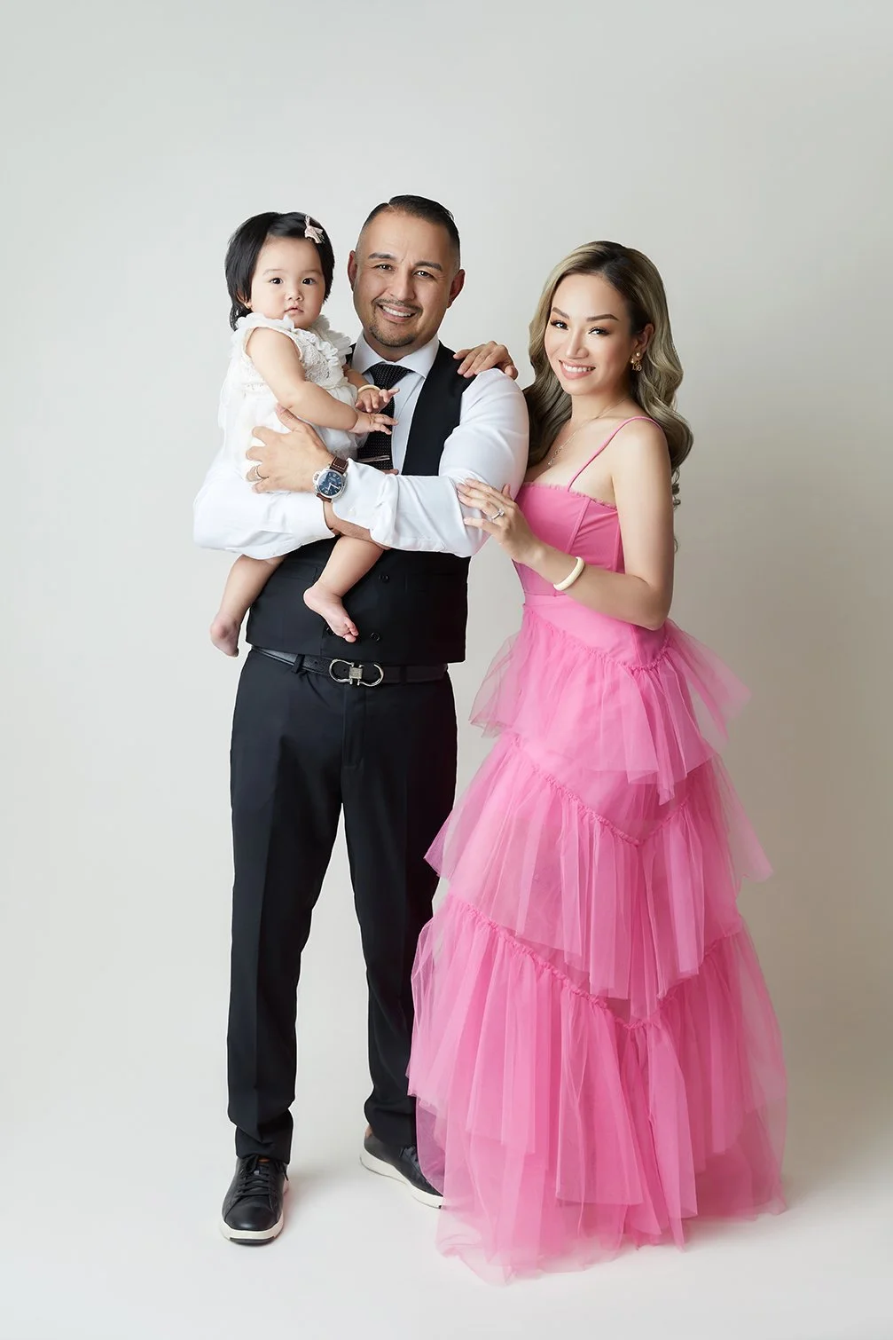 A happy family portrait of a man, woman, and young girl. The man is holding the girl; the woman is standing beside them. The woman is wearing a pink dress, and the man is dressed in a black vest, white shirt, and black pants. The girl is in a white d