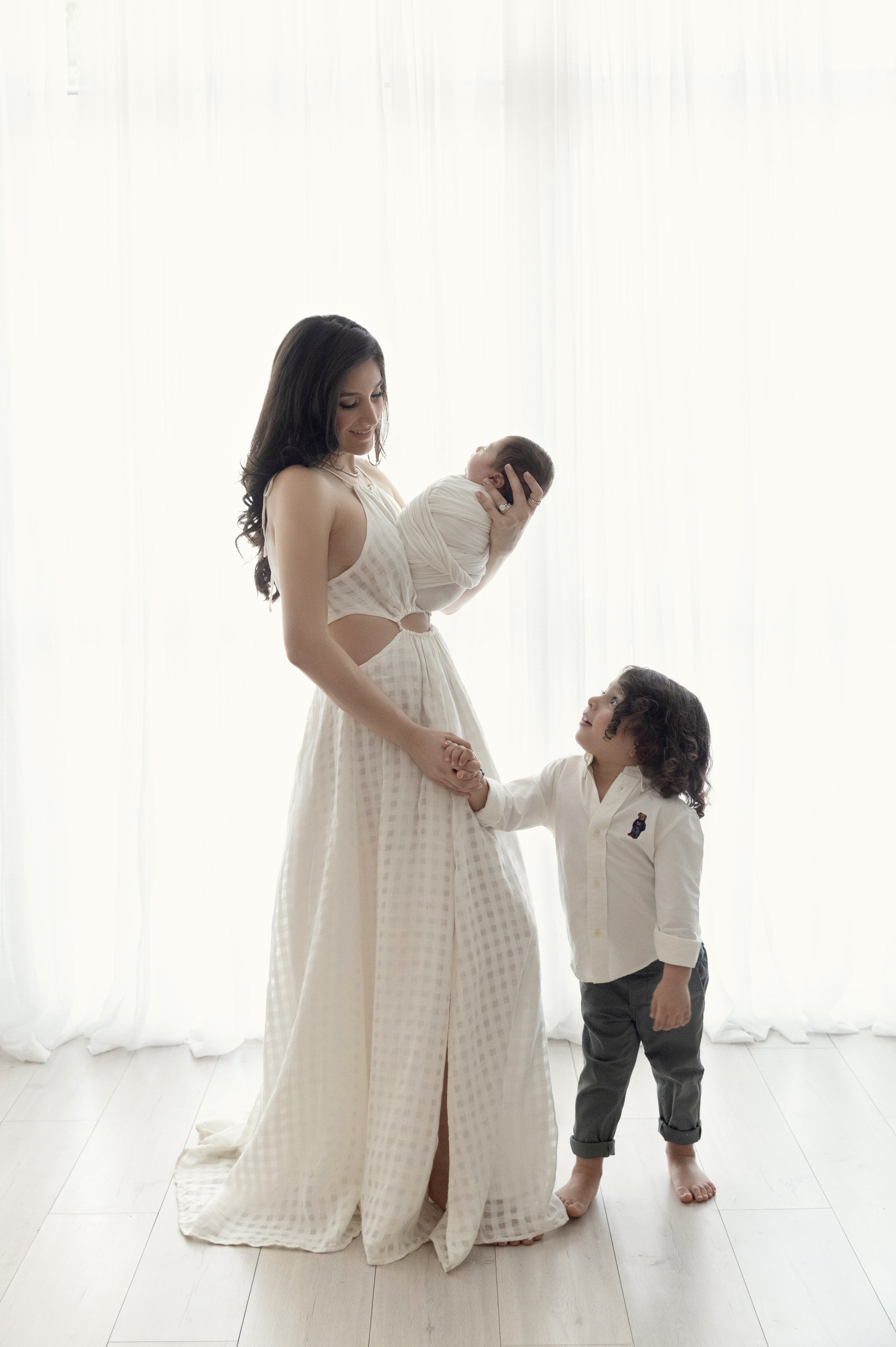 A woman holding a newborn baby and a young child holding her hand, standing in front of a white curtain. The woman wears a light-colored dress, and the young child wears a white shirt and dark pants.