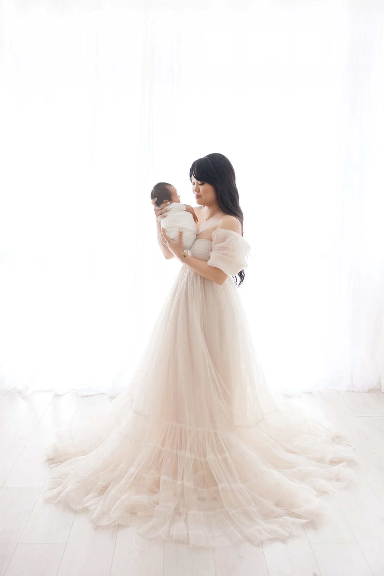 A woman in a flowing, light-colored gown holds a tiny baby wrapped in white cloth, standing on a white wooden floor with bright white curtains in the background.