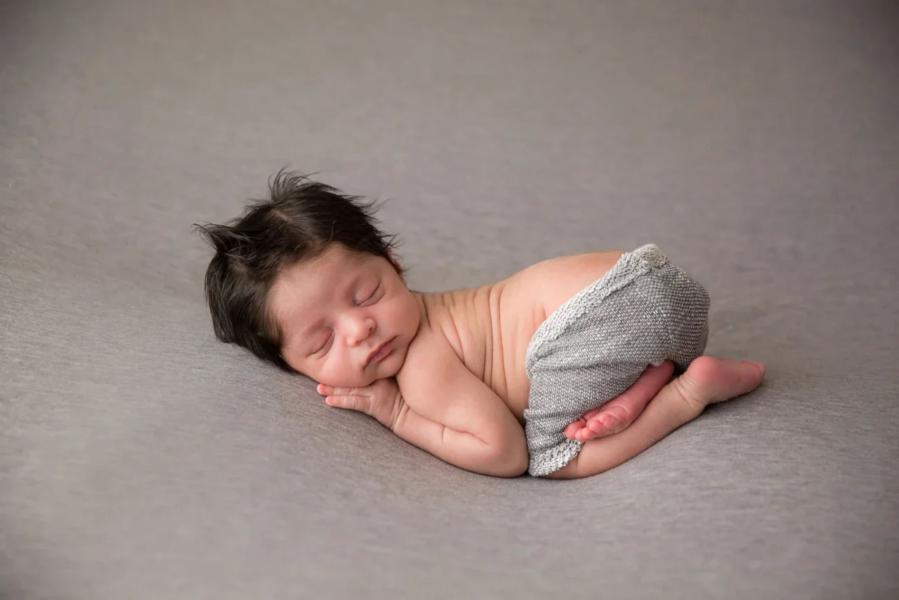 A sleeping baby with dark hair curled up on a gray surface, wearing gray shorts.