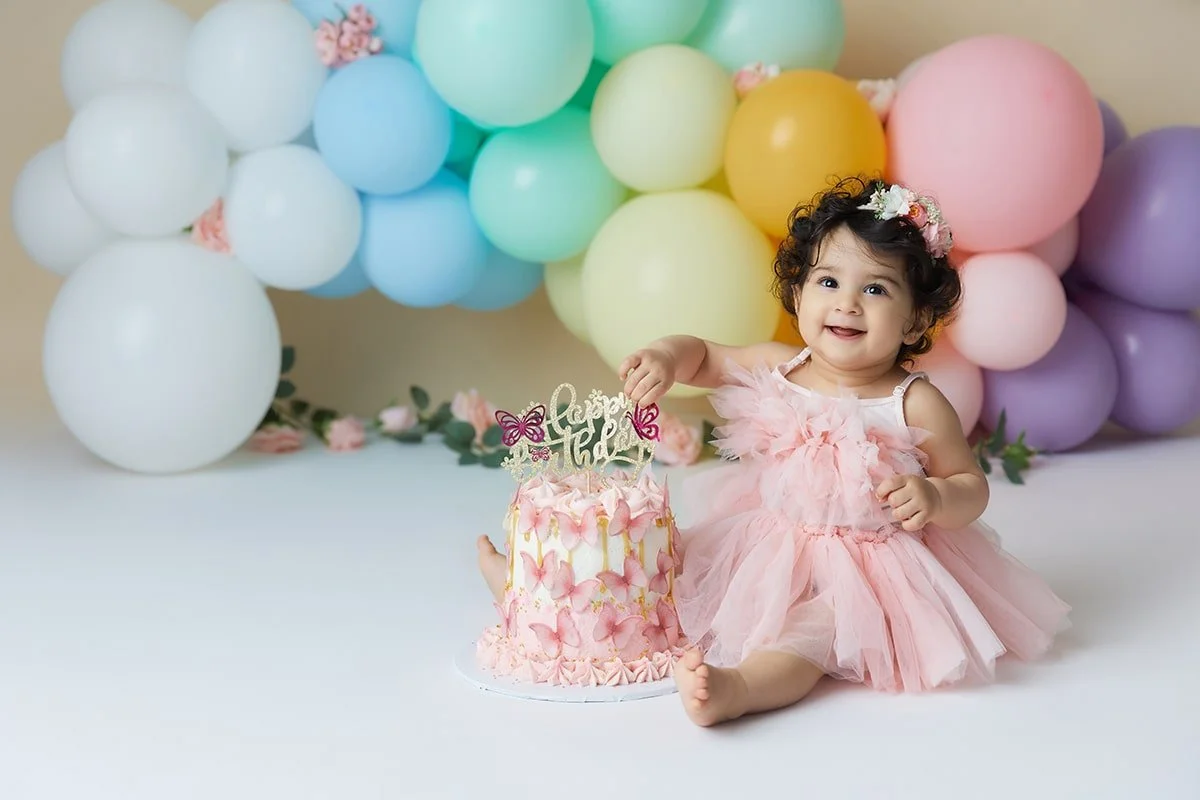 A happy toddler girl in a pink dress sitting on the floor next to a birthday cake decorated with pink butterflies and a topper that says "happy birthday". The background features colorful balloons in pastel shades of white, blue, green, yellow, pink,