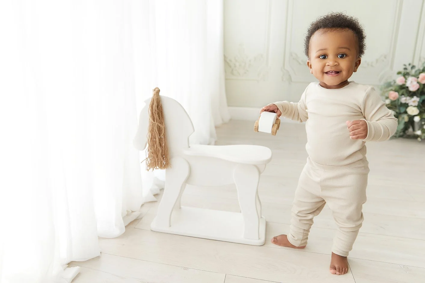 A smiling toddler standing on a light-colored wooden floor, holding a small wooden toy car, with a white decorative chair to the left and a floral arrangement in the background.