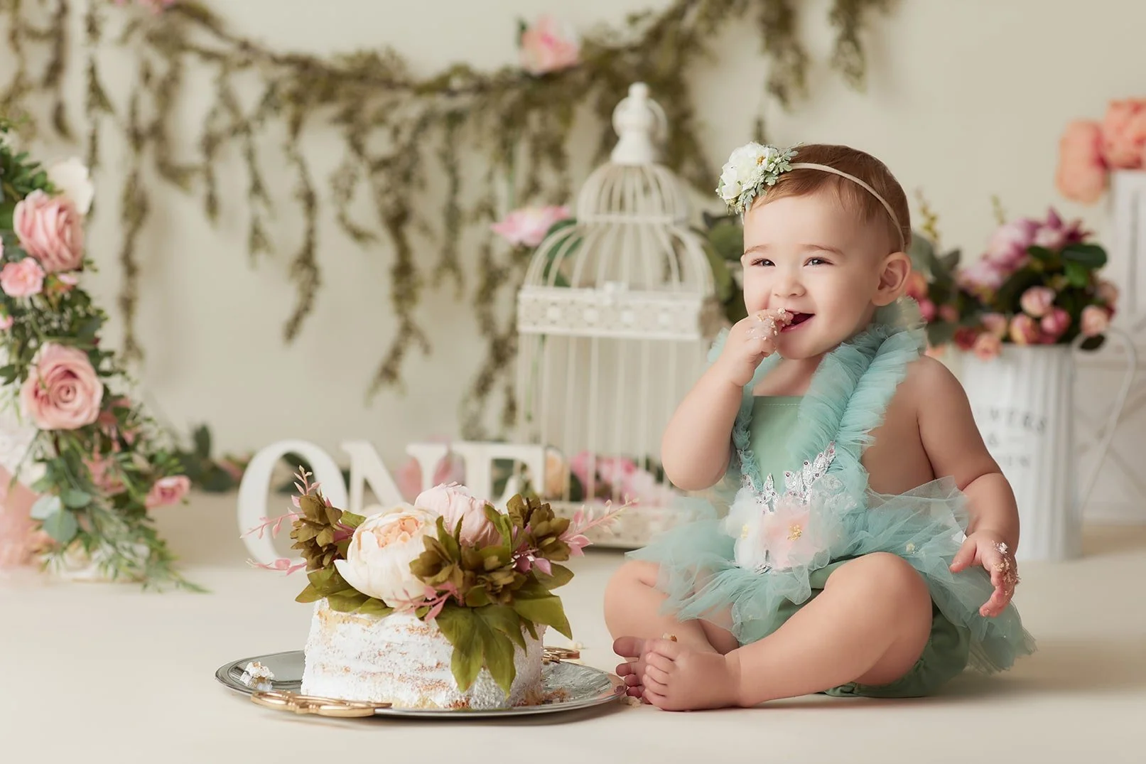 A smiling baby girl in a light blue dress with a floral headband, sitting on the floor with a cake decorated with flowers in front of her, cake crumbs around her mouth and hands, surrounded by pink and white flowers and decorative items.