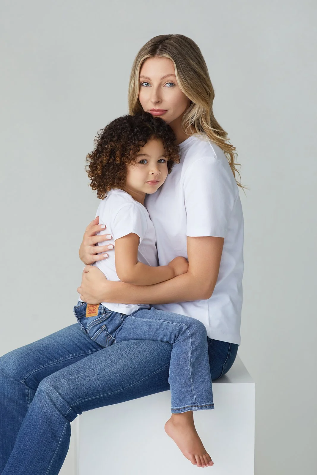 A woman with long blonde hair and a young girl with curly hair sitting together, both wearing white t-shirts and blue jeans, with the girl on the woman's lap and the woman hugging her.
