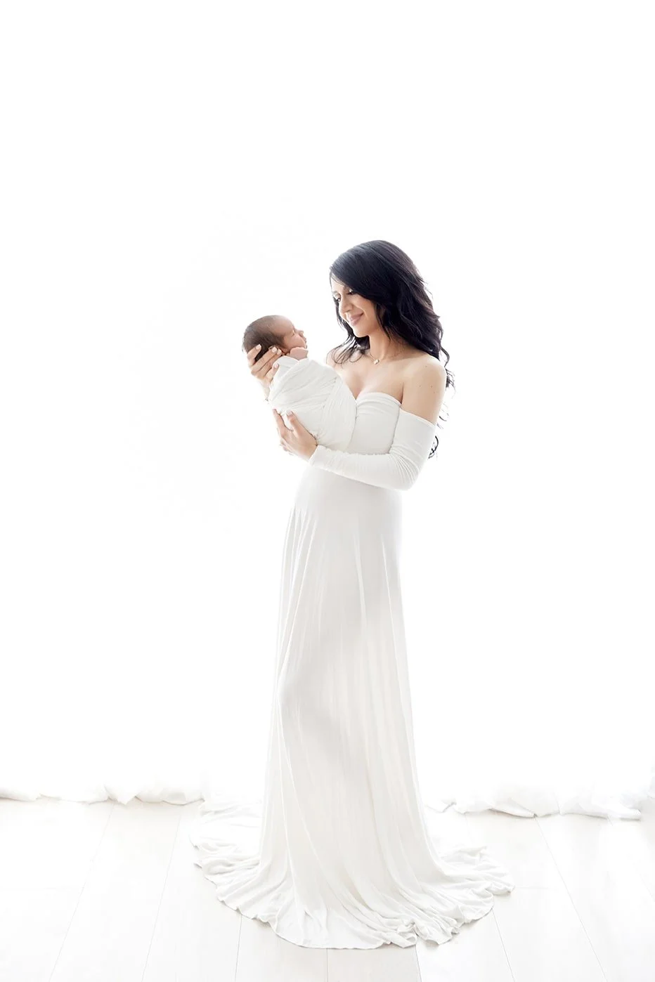 A woman in a white dress holding a baby wrapped in white cloth, standing on a white floor with a bright background.