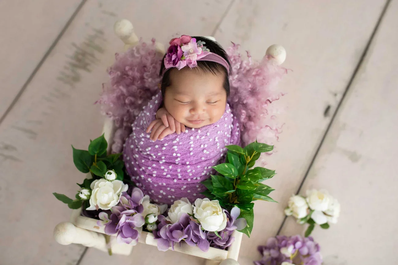 A smiling baby girl with a pink headband and purple dress, lying on a small decorated bed with flowers, surrounded by purple and white flowers on a wooden floor.