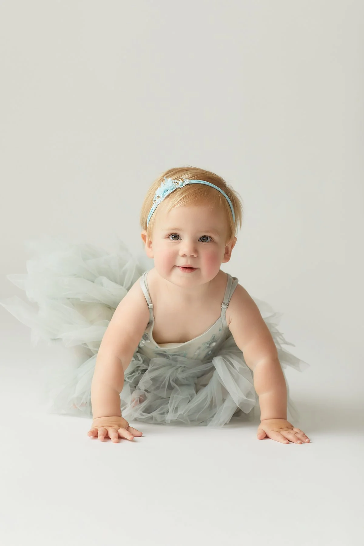 Baby girl crawling on white background, wearing a gray tutu dress and a blue headband.