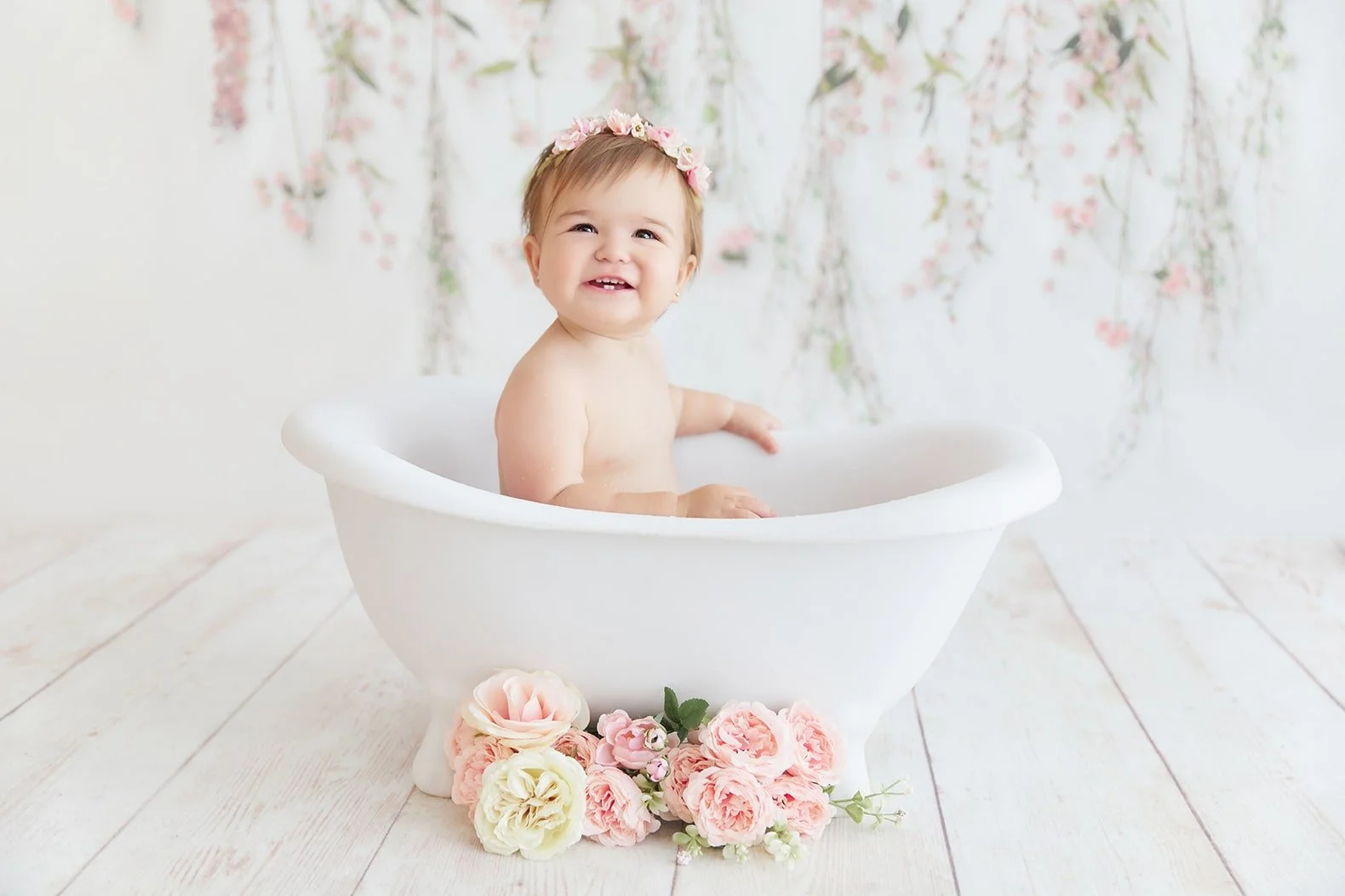 A smiling toddler girl with a floral headband sitting in a white bathtub surrounded by pink roses, with a floral backdrop.