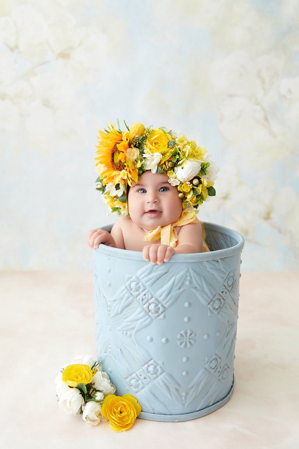 Baby with a large sunflower and yellow flower crown, sitting in a decorative gray bucket, with yellow and white flowers on the table