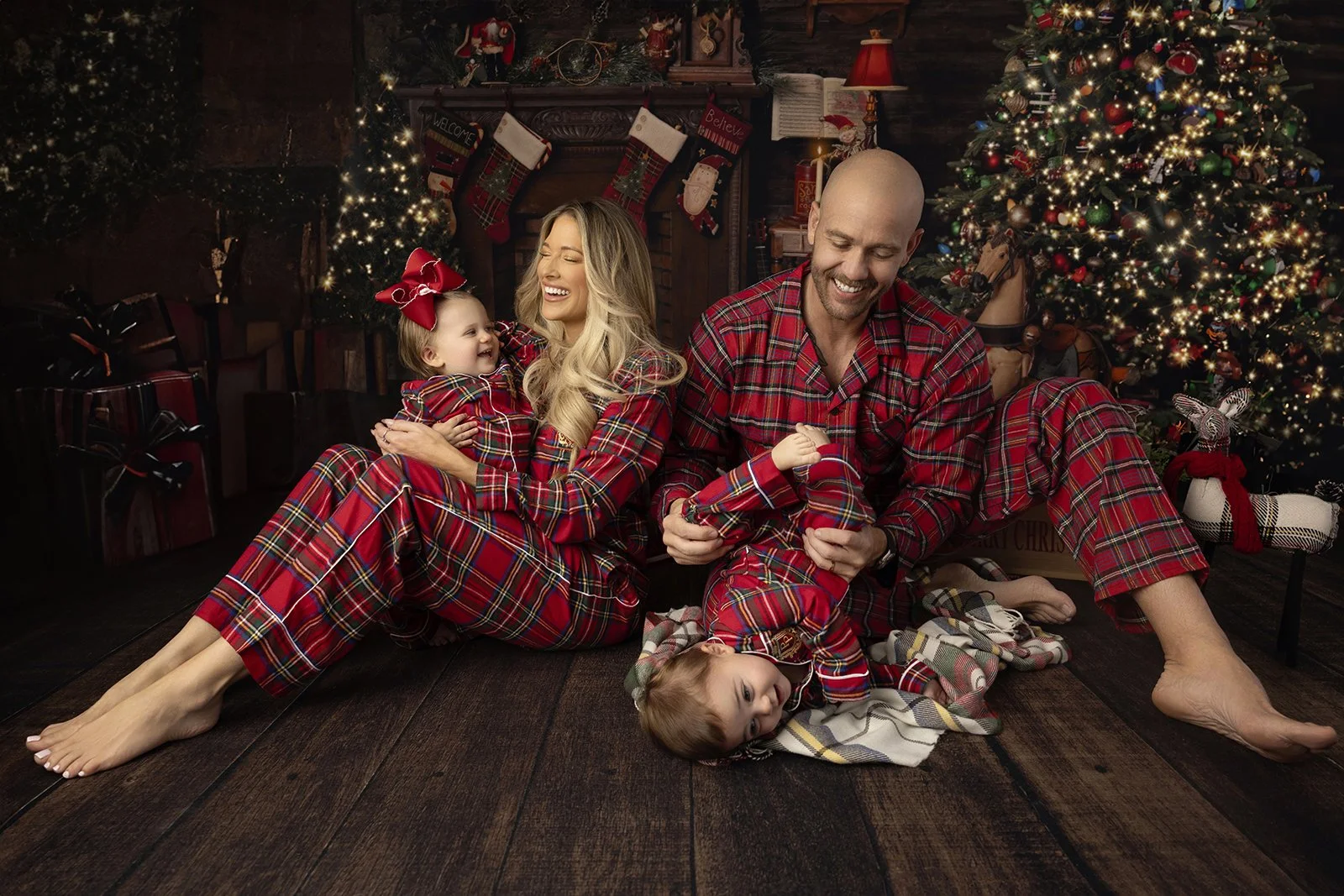 Family of four wearing matching red plaid pajamas sitting on a wooden floor in front of a decorated Christmas tree, engaging in playful playtime and sharing joyful moments.