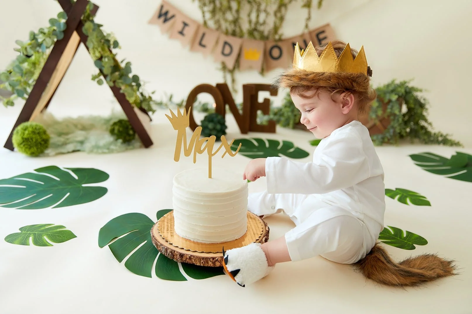 A young boy dressed as a fox, wearing a golden paper crown, sitting on a jungle-themed party table, celebrating his birthday with a white frosted cake that has a gold 'Max' cake topper. The background features green tropical leaves, a wooden teepee d