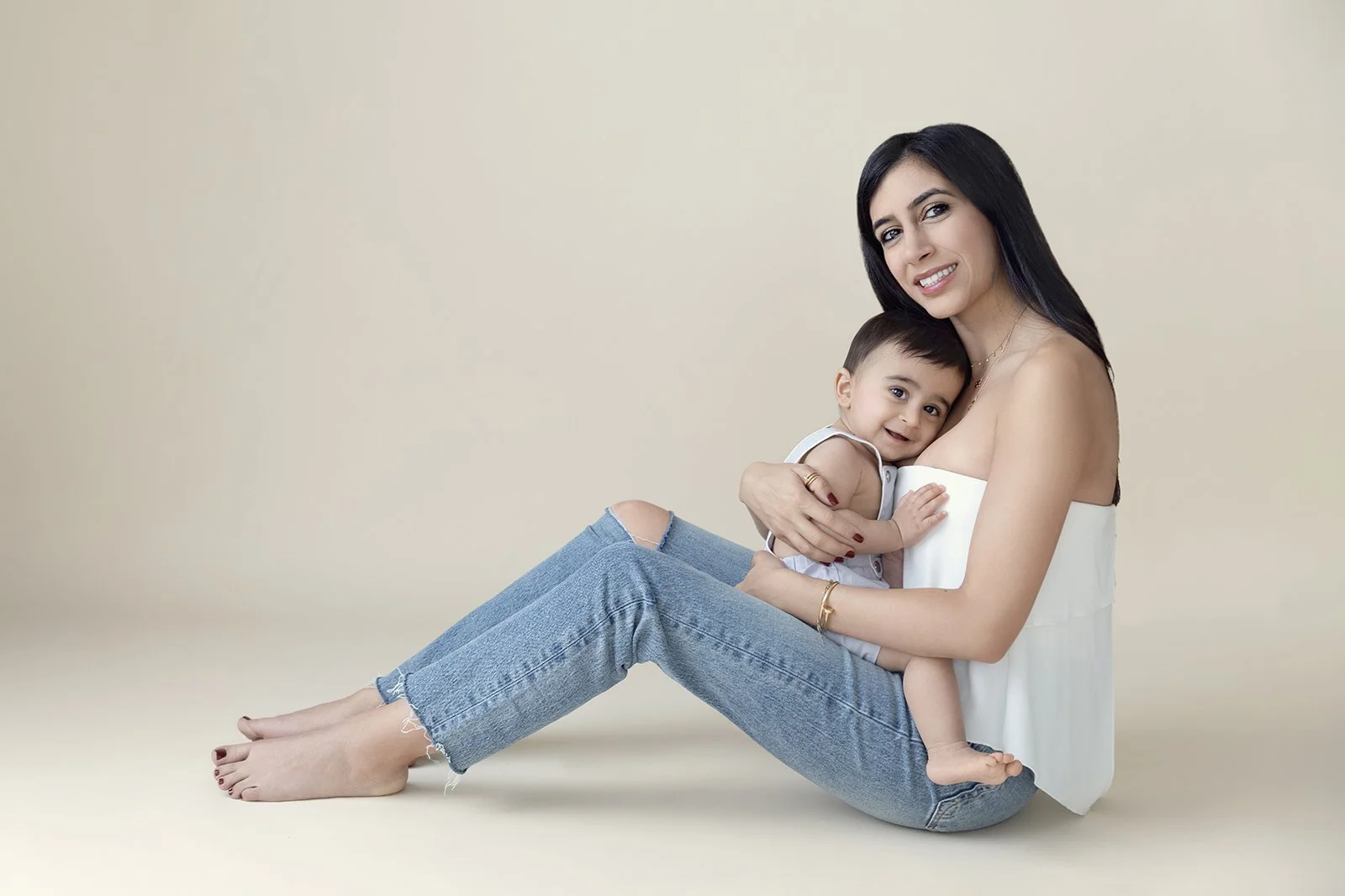A woman sitting on the floor hugging a young child, both smiling at the camera against a neutral background.