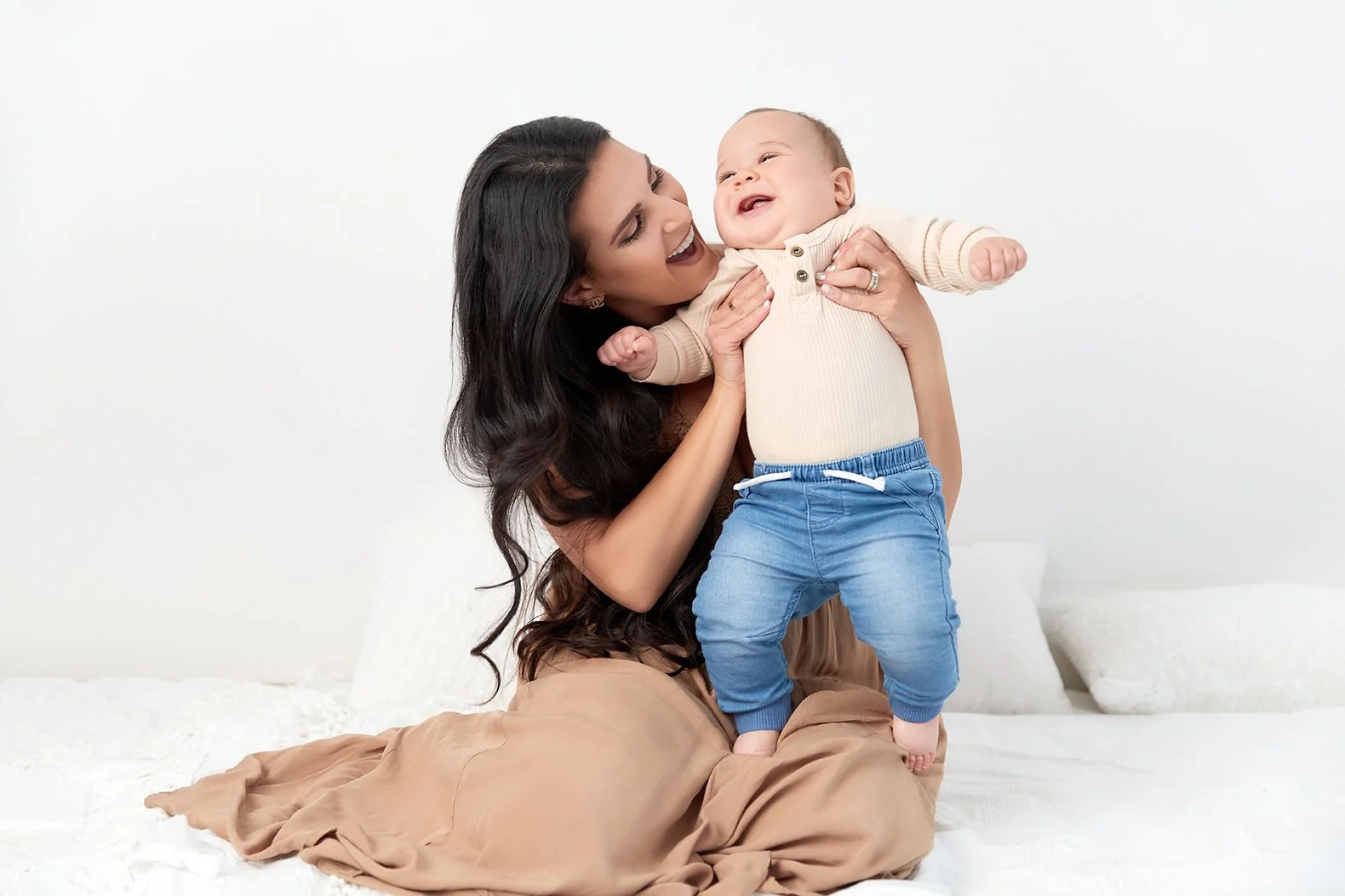 A woman with long dark hair holding a smiling baby boy with light skin, dressed in a beige sweater and blue pants, as they share a joyful moment on a white bed.