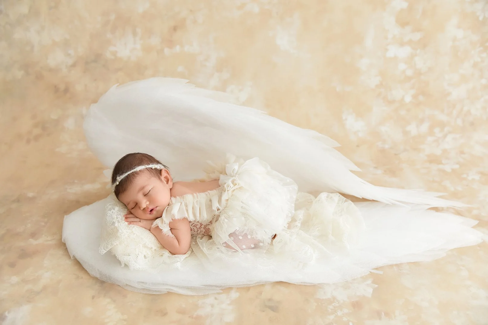 Sleeping baby girl dressed in white lace, lying on a white feathered angel wing-shaped prop, against a beige background.