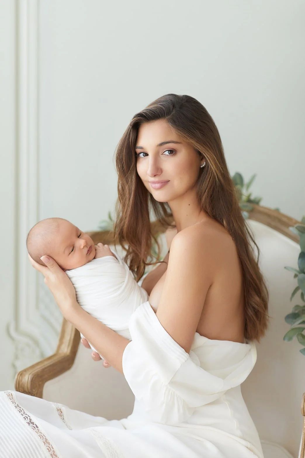 A woman holding a sleeping newborn baby in her arms while sitting on a vintage-style wooden bench with white upholstery, decorated with green foliage, in a bright room.