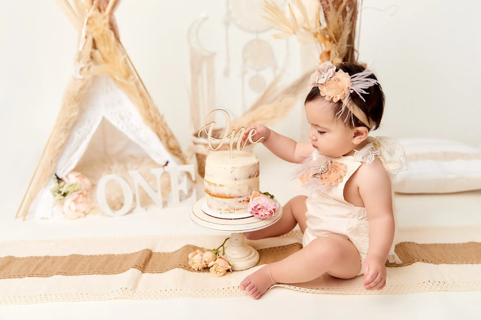 A baby girl celebrating her first birthday sitting on a blanket with a small cake decorated with a 'One' topper, surrounded by pink flowers and a rustic background with a teepee and 'ONE' sign.