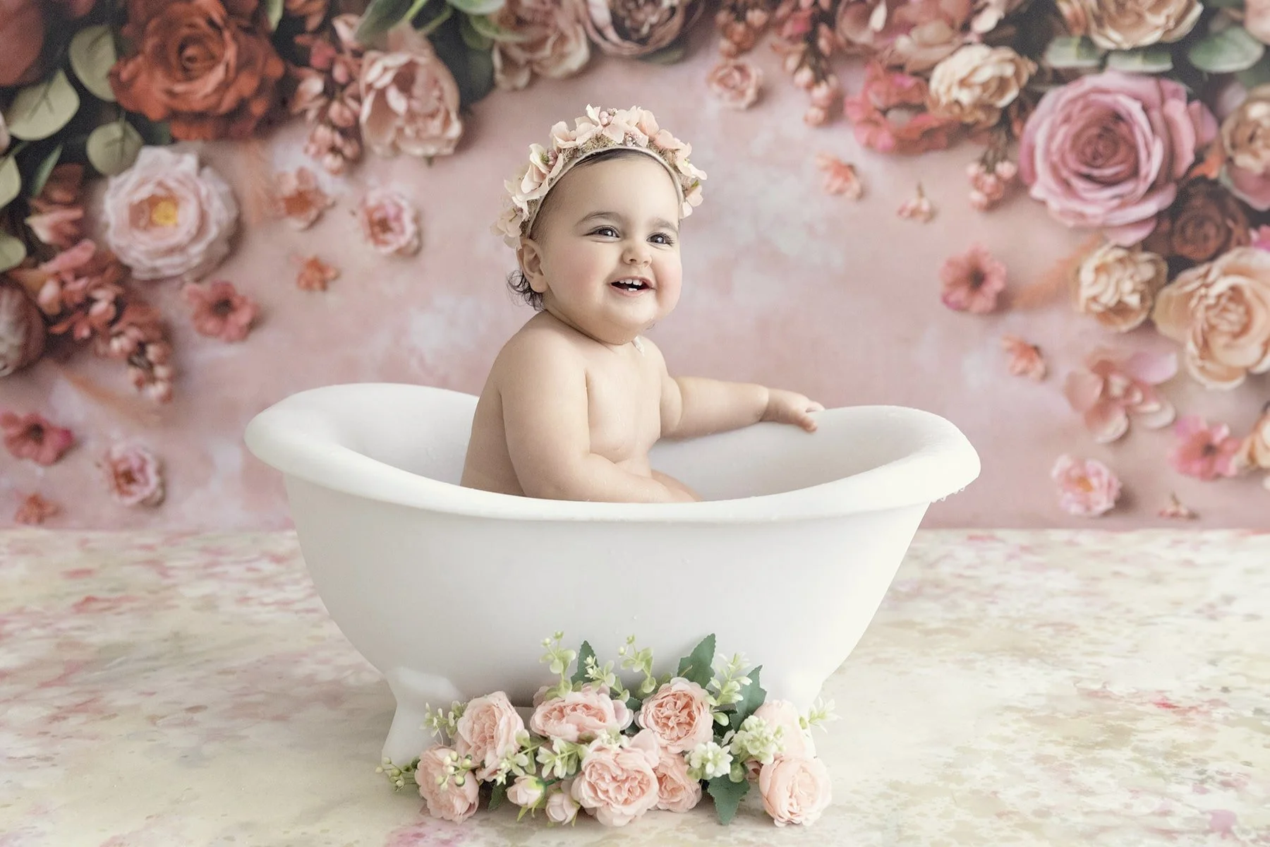 A smiling baby girl with a flower crown sitting in a white bath tub decorated with pink and white flowers, against a pink floral backdrop.