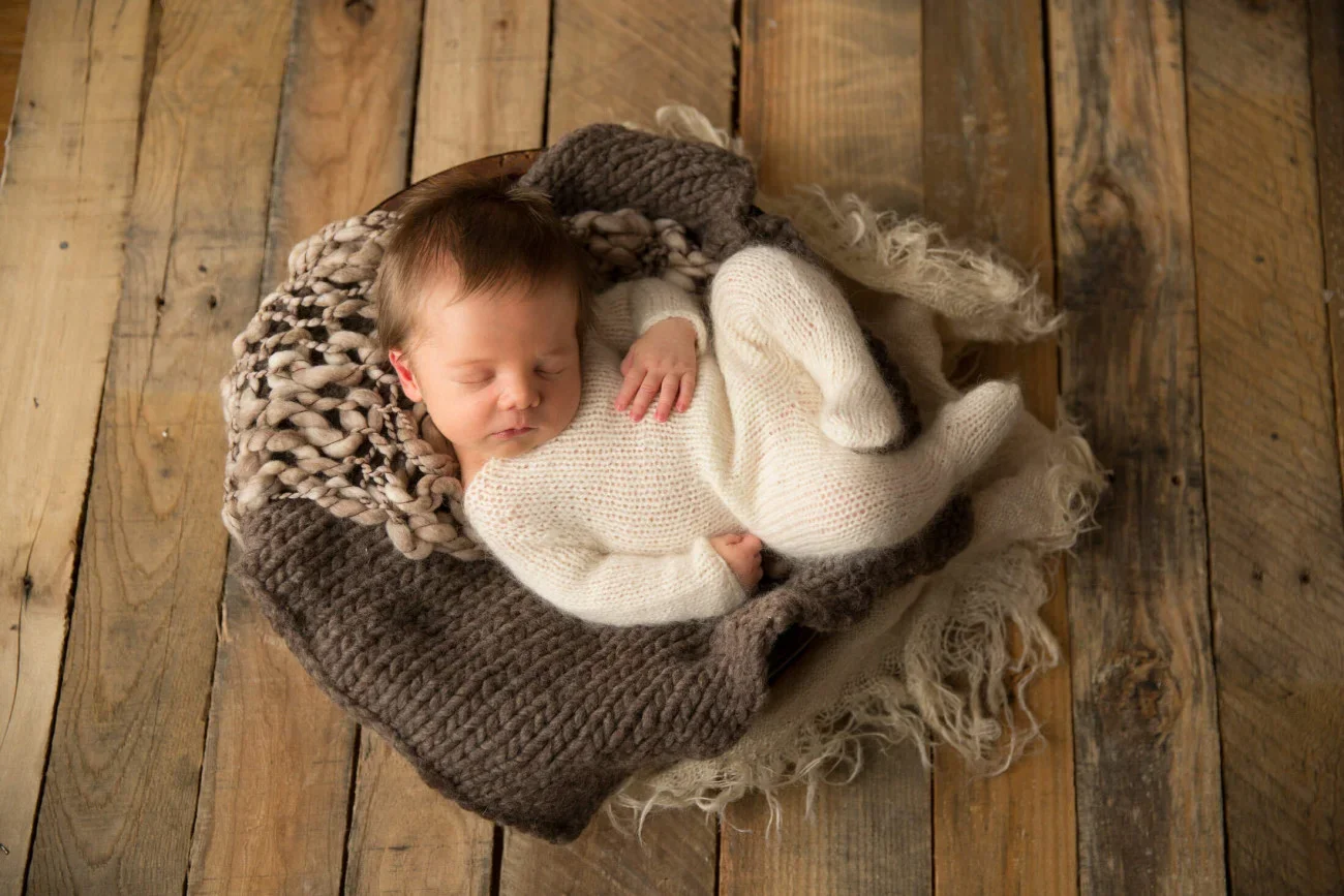 A sleeping baby wrapped in a white knit outfit, lying on a soft, chunky knit blanket inside a woven basket, on a wooden floor.