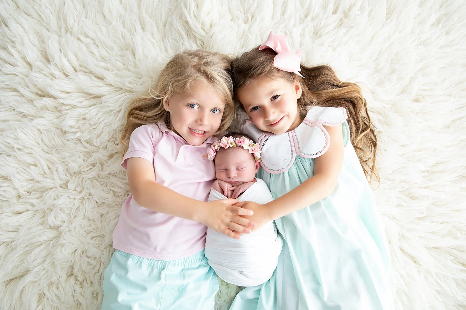 Two young girls and a newborn baby girl lying on a fluffy white rug, smiling and embracing each other. The newborn wears a floral headband, the girl on the left has blonde hair and a pink shirt, the girl on the right has brown hair, a pink bow, and a
