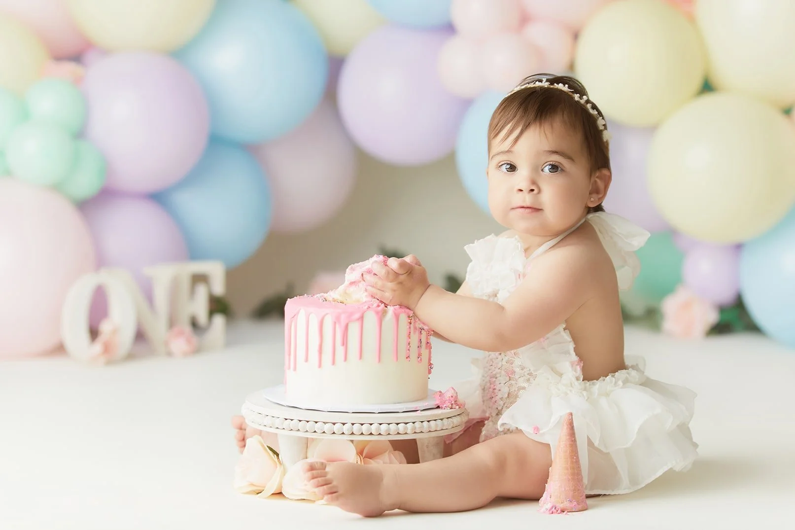 A young girl in a white dress celebrating her first birthday with a pink and white cake, surrounded by pastel balloons and decorative decorations.