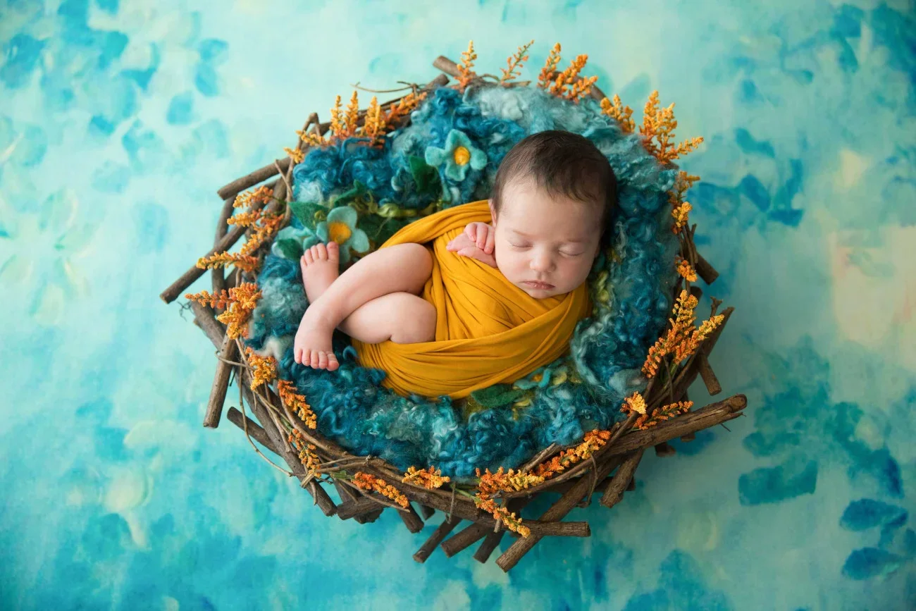 A sleeping baby wrapped in a yellow cloth, lying in a nest of blue and green textured fabric, surrounded by yellow-orange faux flowers, against a watercolor blue-green background.