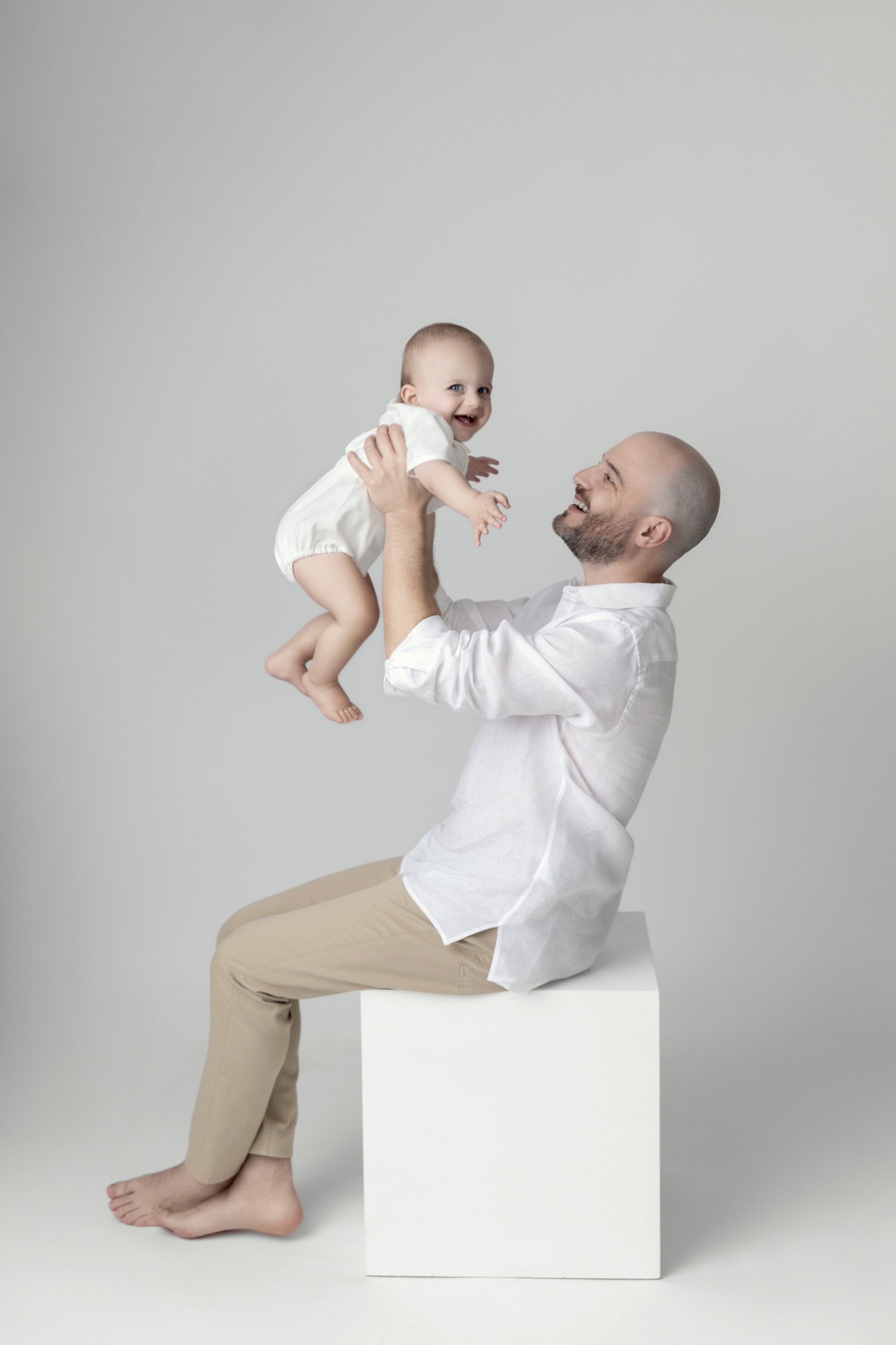 A man with a beard sitting on a white cube, holding a smiling baby girl in a white outfit in the air, both appearing joyful.