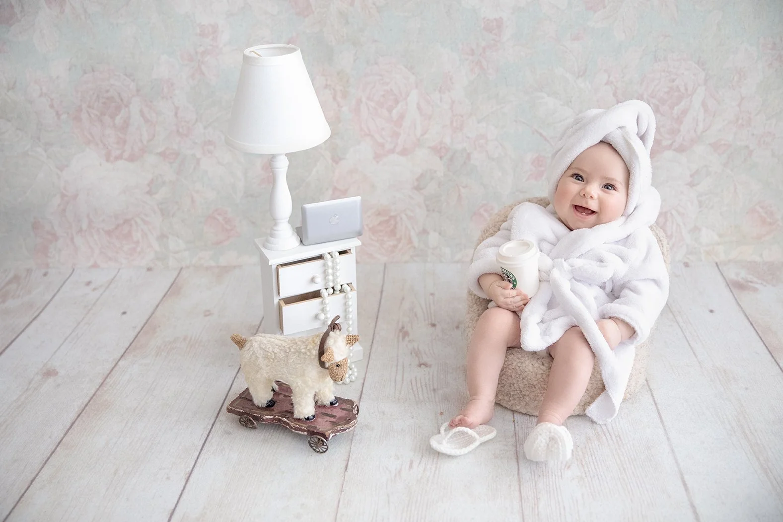 A smiling baby wearing a white bathrobe and towel hat, sitting on a small beige chair holding a cup, with a cozy homemade stuffed sheep on a toy cart, a white lamp, and a small table with a smartphone on it, in front of a pastel floral wallpaper and white wooden floor.