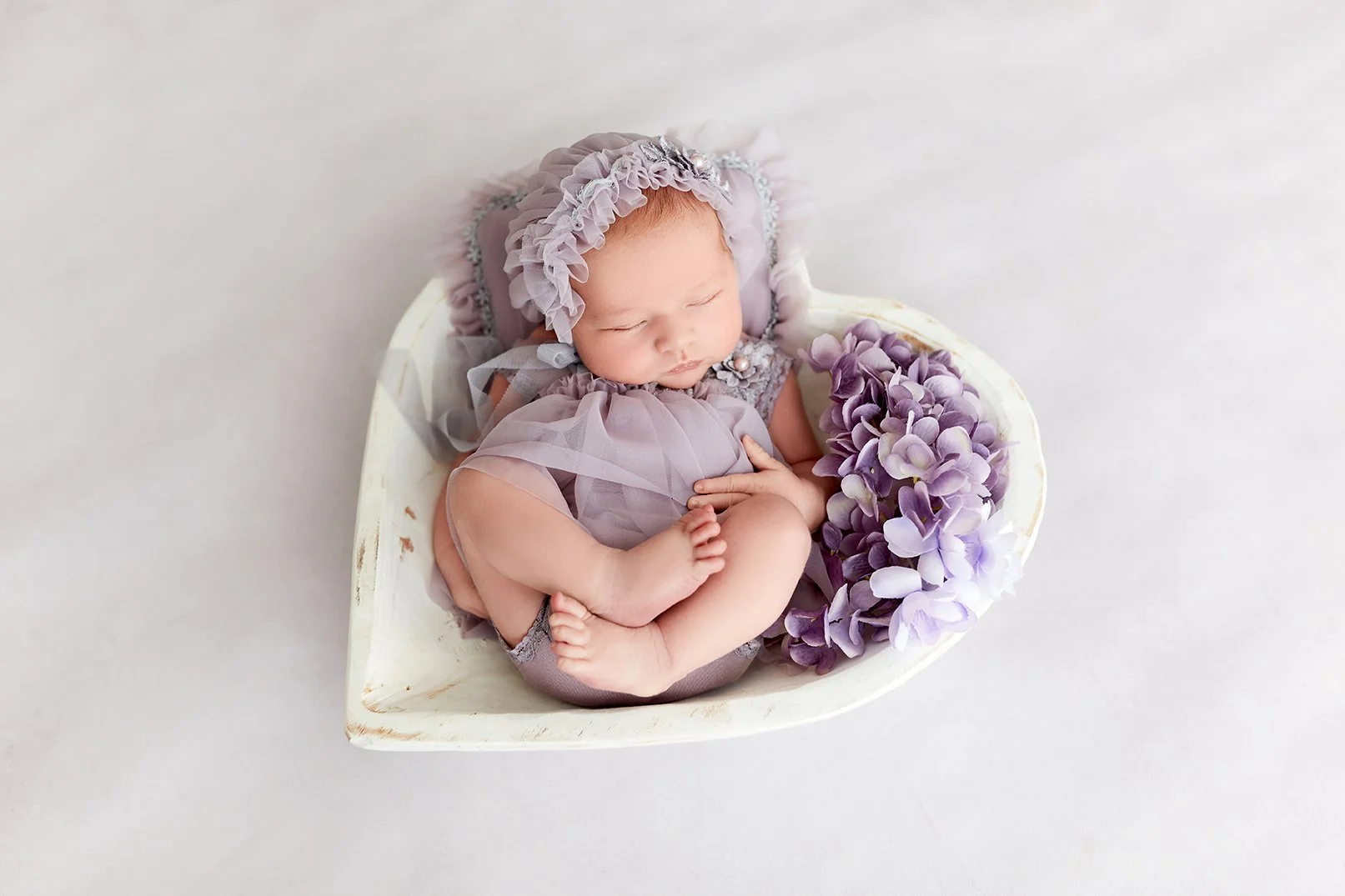 A sleeping baby girl in purple clothes and a bonnet, curled up in a white bowl, with purple hydrangea flowers beside her.