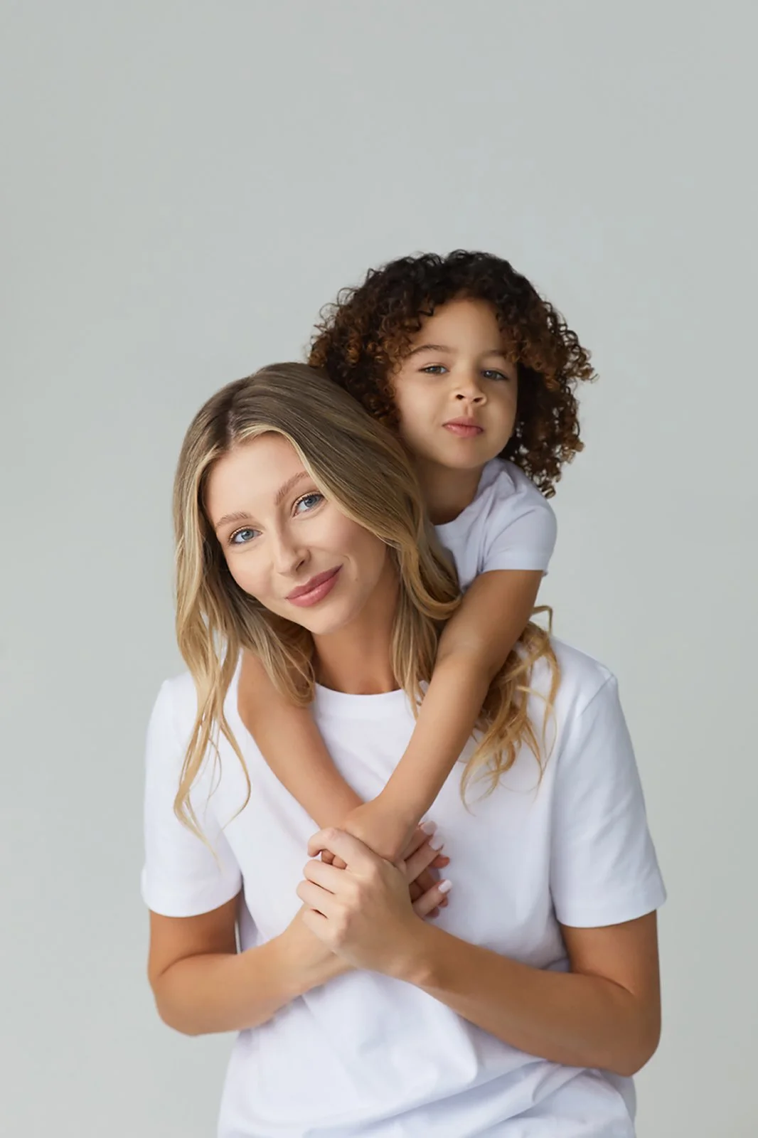A woman with blonde hair and a young girl with curly hair are together. The girl is on her shoulders, and both are wearing white shirts. They are looking at the camera with a neutral expression. The background is plain and light-colored.