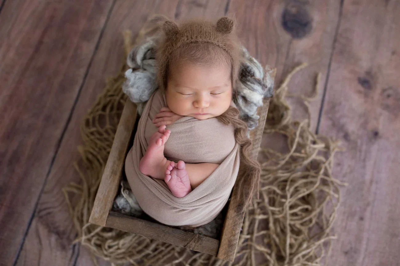 A sleeping baby wrapped in beige cloth, lying in a small wooden bed with wool and fur accents, on a rustic wooden floor surrounded by straw.