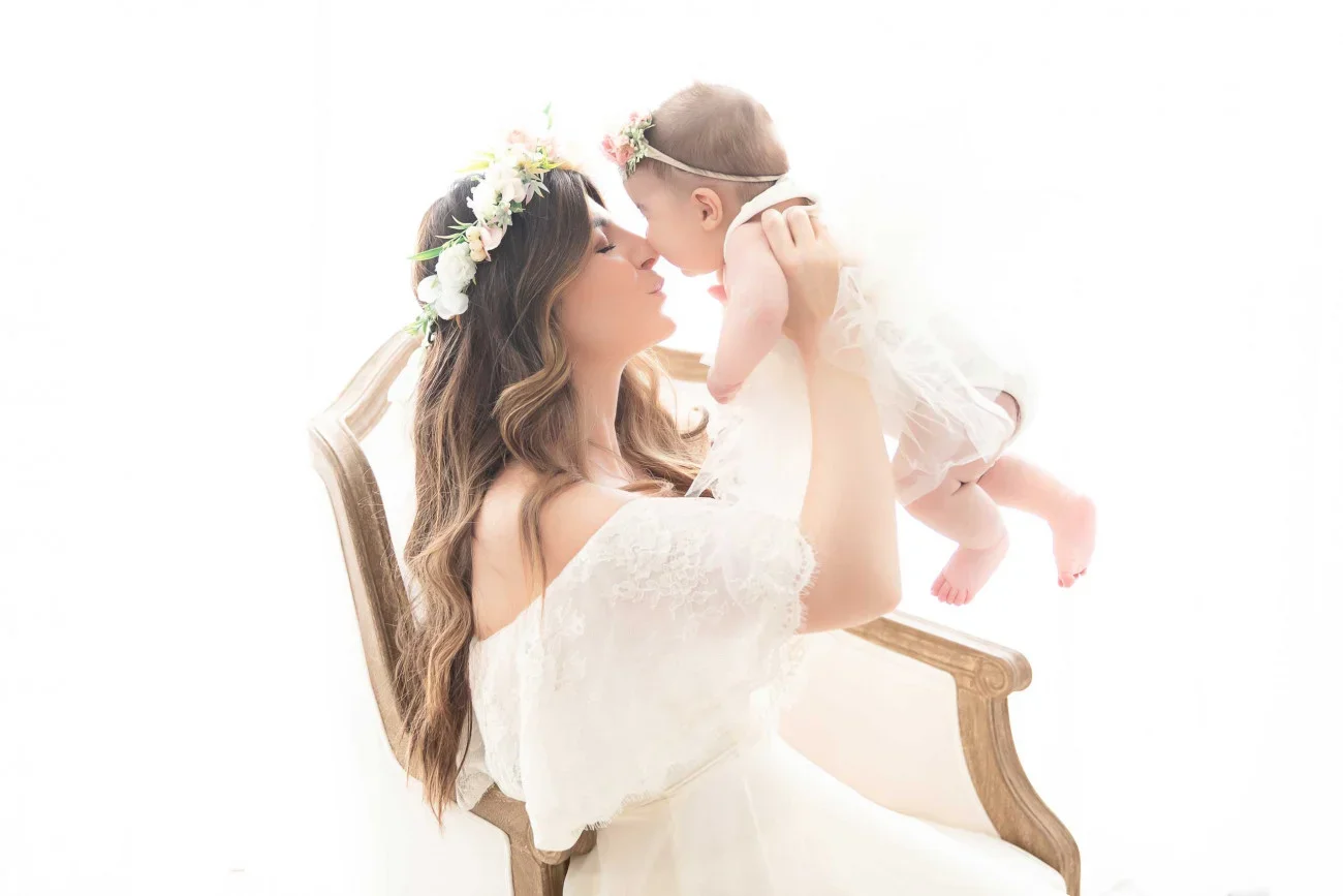 A woman with long, wavy brown hair wearing a white dress and a floral crown holds a baby girl in her arms. The baby is dressed in a light-colored dress with a flower headband, and they are touching noses in a tender moment, with a white background.