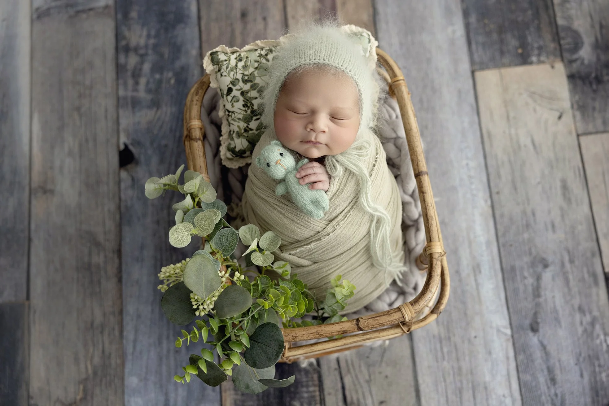 A sleeping newborn baby wrapped in beige cloth, lying in a small wicker basket with a floral pillow, holding a small green stuffed duck, surrounded by green eucalyptus leaves, on a wooden floor.