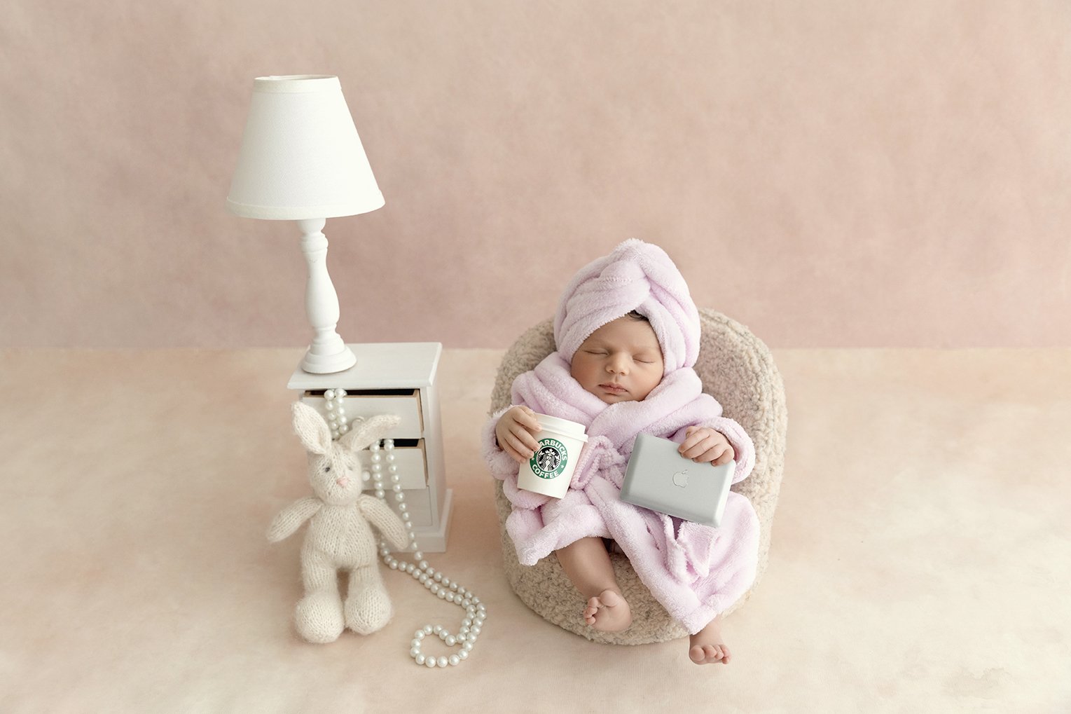 A baby dressed in a pink towel and towel wrapped on her head, sitting in a small chair, holding a Starbucks coffee cup and a silver laptop, surrounded by soft decor including a lamp, a white knit bunny toy with pearl necklaces, set against a pink bac