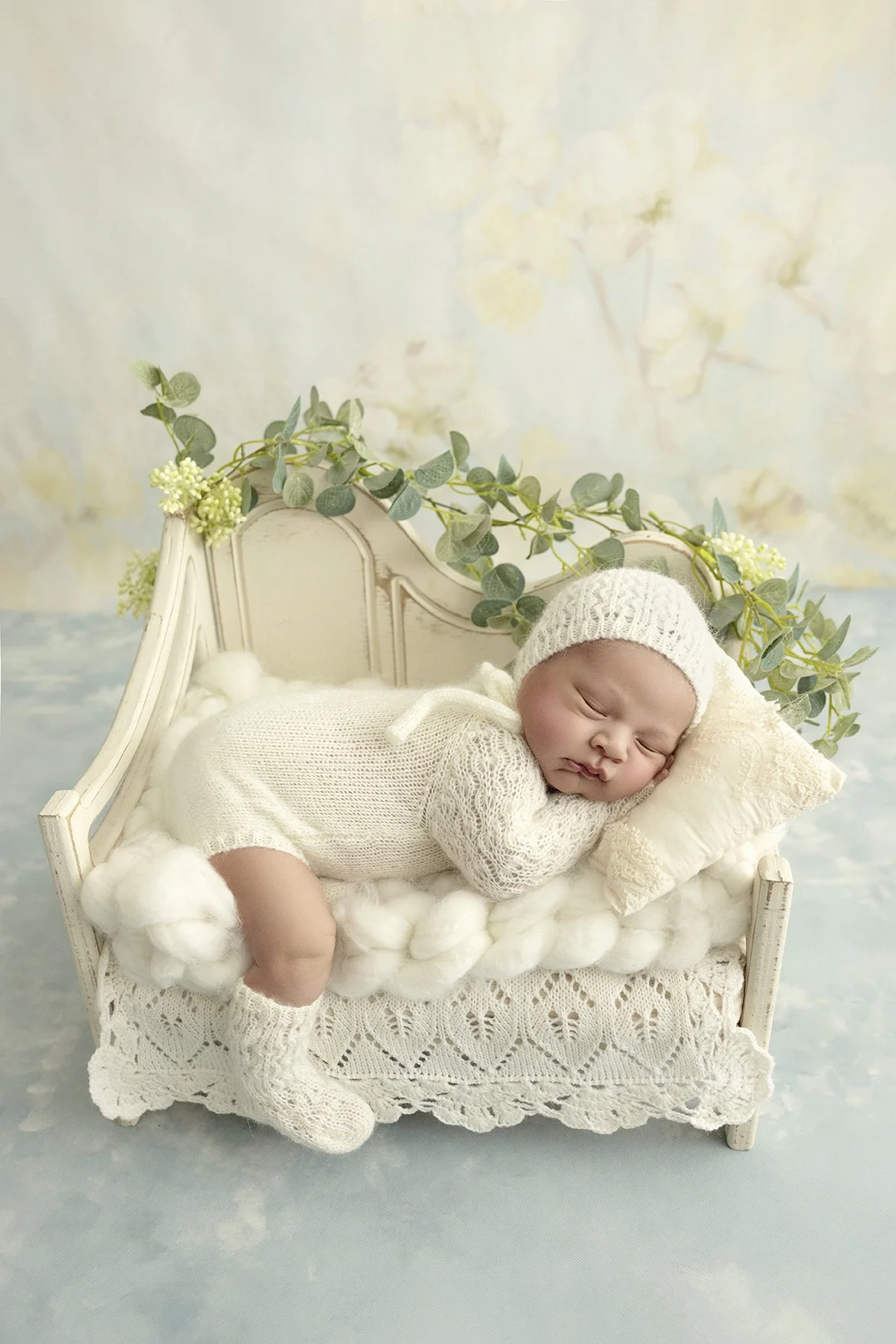 A sleeping baby in a white knit outfit and hat, lying on a miniature white bed with plush bedding, surrounded by greenery and soft pastel background.