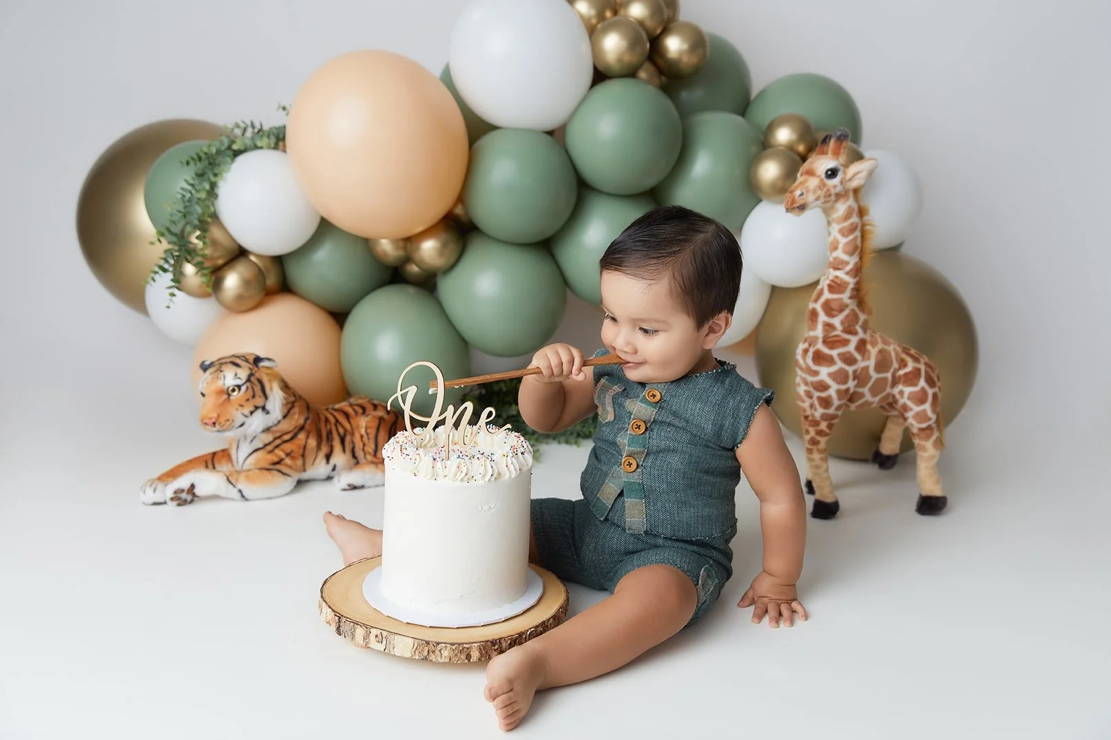 A young boy in a dark green sleeveless romper celebrating his first birthday with a cake inscribed with 'One', surrounded by plush tiger and giraffe toys, with a backdrop of green, white, and gold balloons.