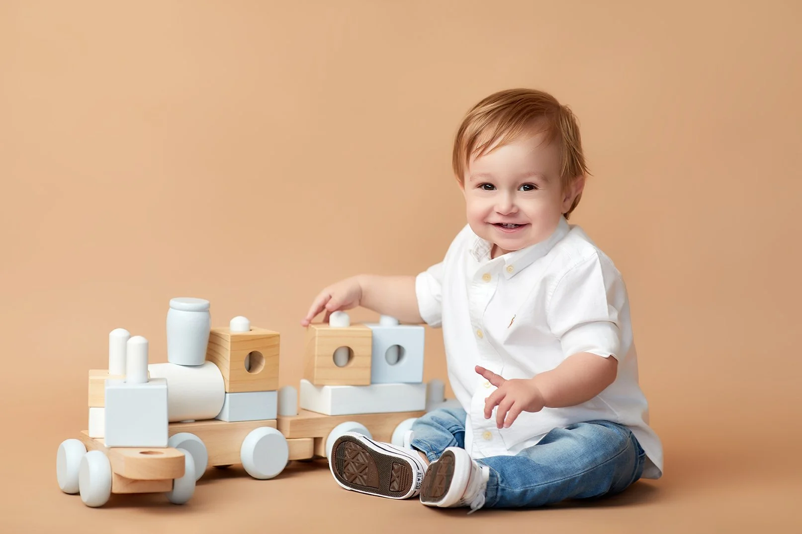 Baby boy sitting on the floor playing with a wooden toy train, wearing a white shirt and jeans, with a peach-colored background.