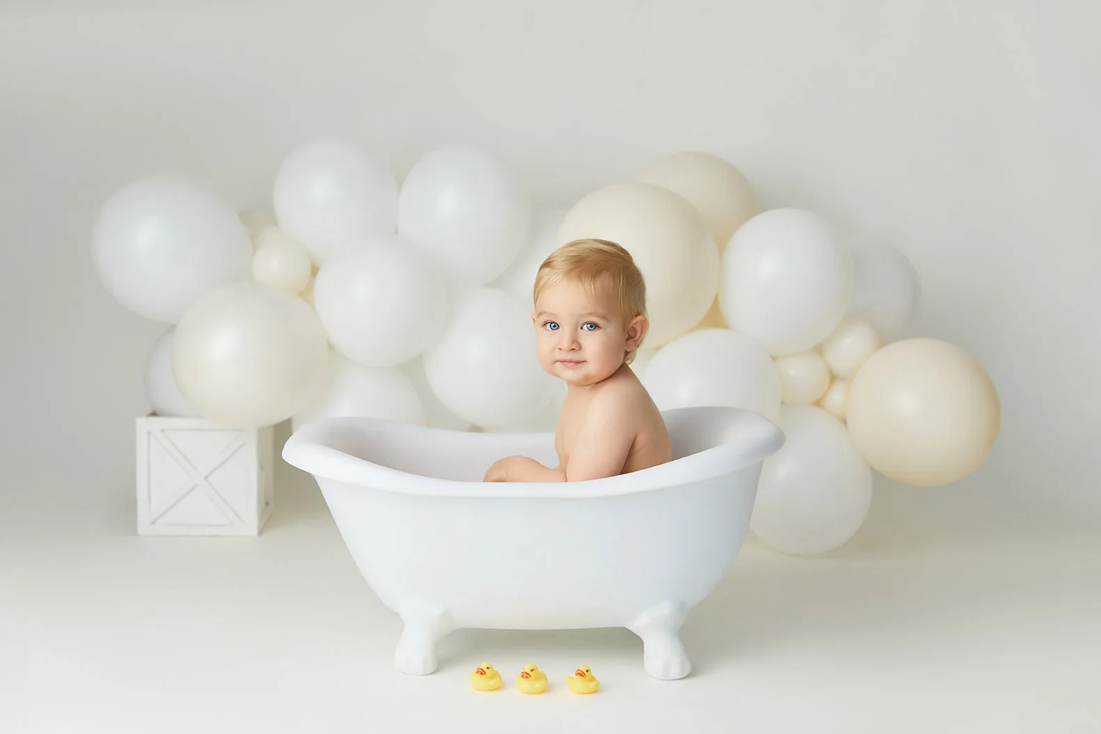 A young child with blonde hair and blue eyes sitting in a white bathtub with a backdrop of white balloons and three small rubber ducks in front, on white background.