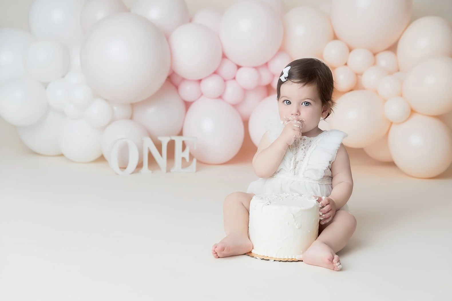 A young girl with a white dress and a small white bow in her short dark hair sitting on the floor during her first birthday celebration, eating cake with pink and white balloons in the background and a white layered cake in front of her.