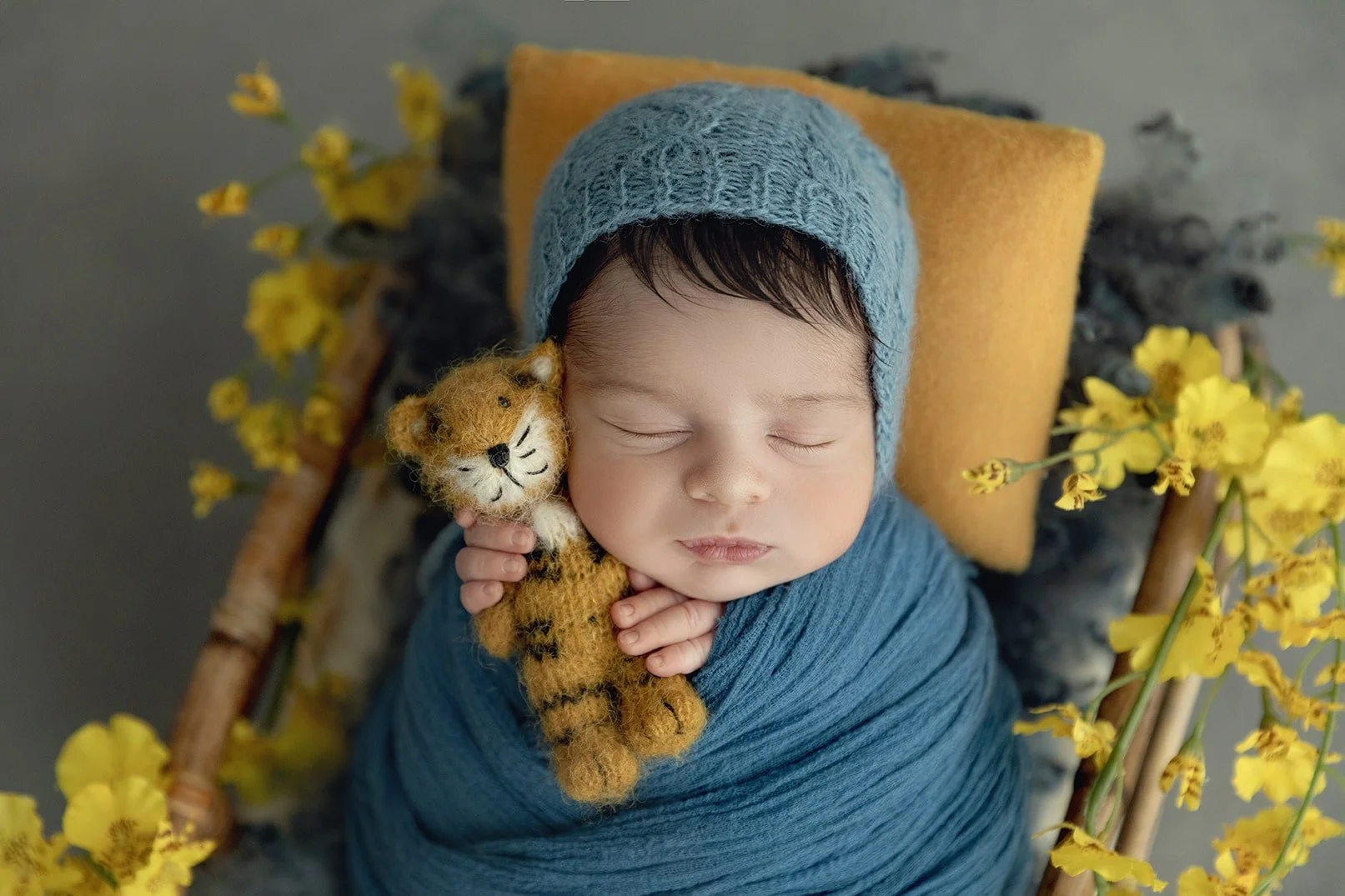 A sleeping baby dressed in a blue knit hat and wrapped in a blue blanket, holding a small felt tiger stuffed animal, surrounded by yellow flowers on a wooden cradle.